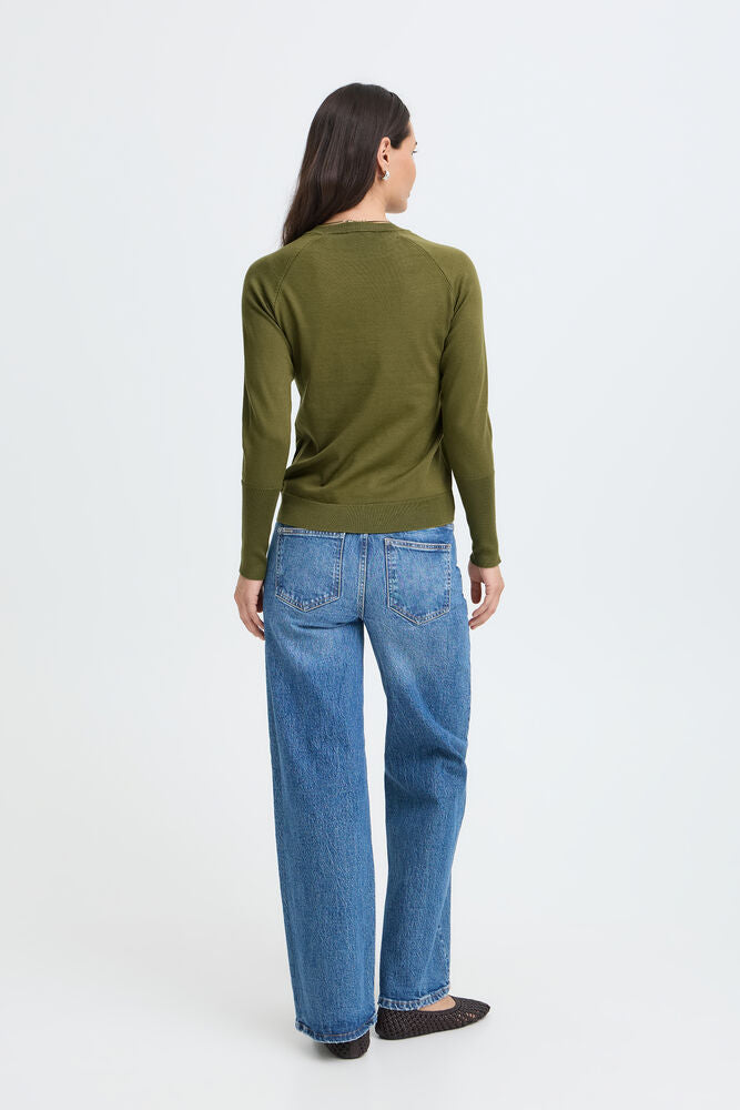 A woman viewed from the back wearing an olive green long-sleeve top and blue jeans against a plain light gray studio background.