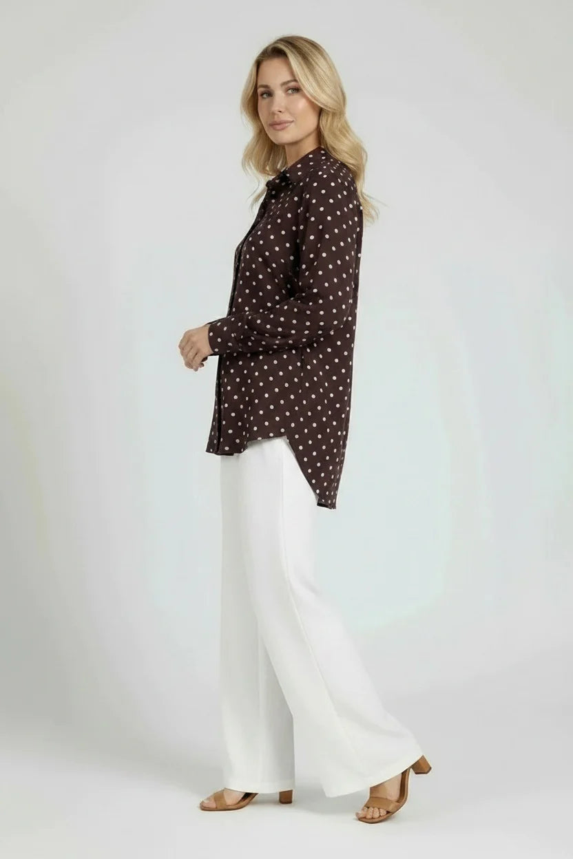Woman wearing a dark patterned blouse and white pants on a plain background