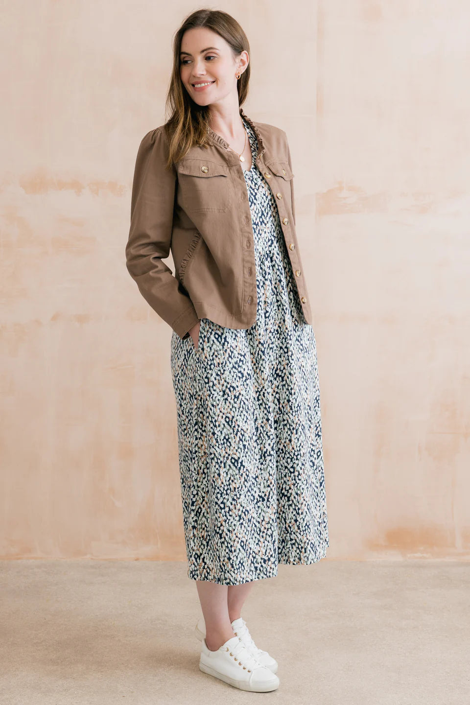Woman wearing a floral dress, brown jacket, and white sneakers against a beige background