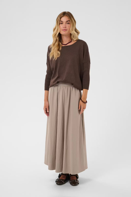 A woman modeling a dark brown long-sleeve top and a light beige maxi skirt against a plain light gray studio background.