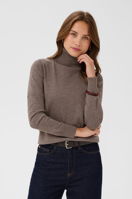 A woman wearing a taupe brown ribbed turtleneck sweater and dark blue jeans, posing against a light gray studio background.