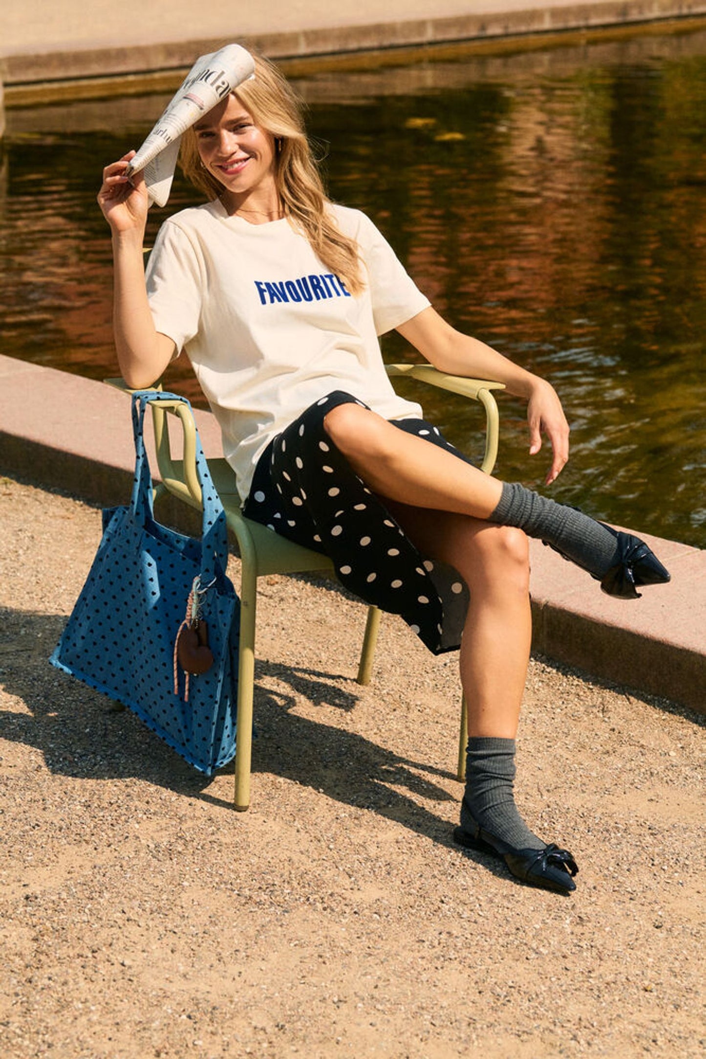 Woman sitting by a pond holding a blue bag, wearing a white t-shirt with text and dark polka dot skirt.