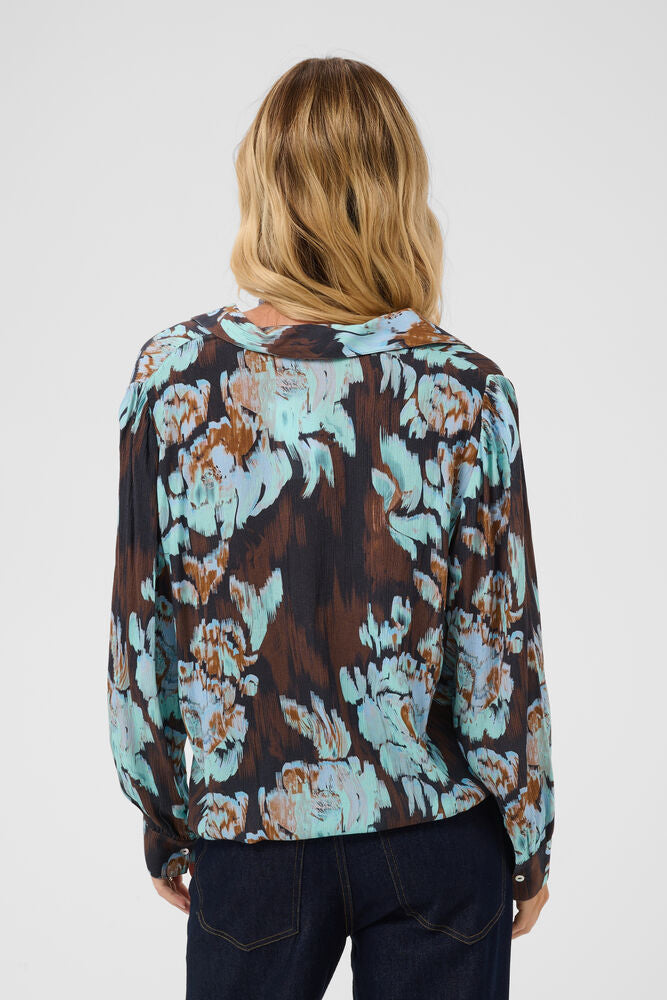 A woman viewed from behind wearing a long-sleeve blouse with a brown, light blue, and beige floral pattern, paired with dark blue jeans, against a light gray studio background.