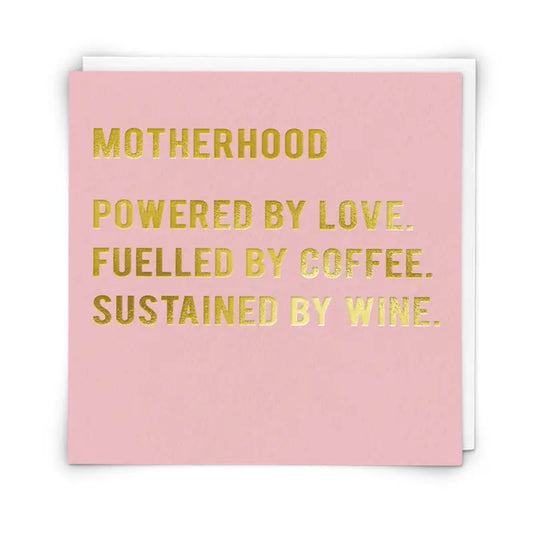 A pink greetings card with gold foiled text that reads: 'MOTHERHOOD Powered by love. Fuelled by coffee. Sustained by wine.'