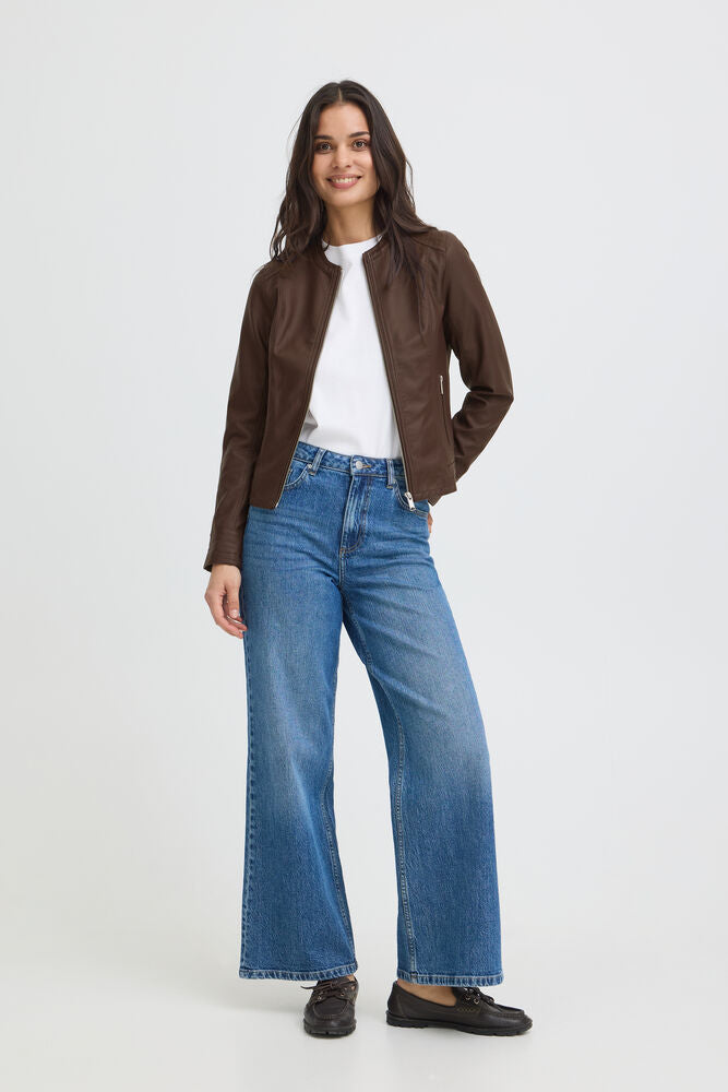 A woman modeling a dark brown suede moto jacket, styled with a white crewneck t-shirt, wide-leg blue denim jeans, and dark brown leather loafers, against a plain light gray studio background.