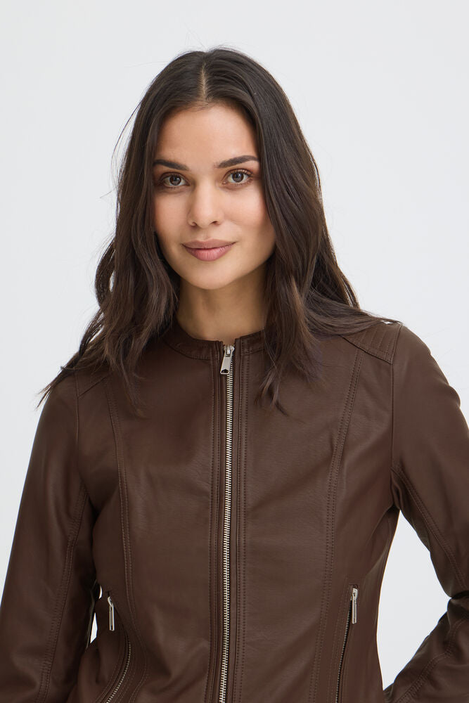 A woman with long dark brown hair wearing a brown faux-leather jacket with silver zippers, photographed against a light gray studio background.