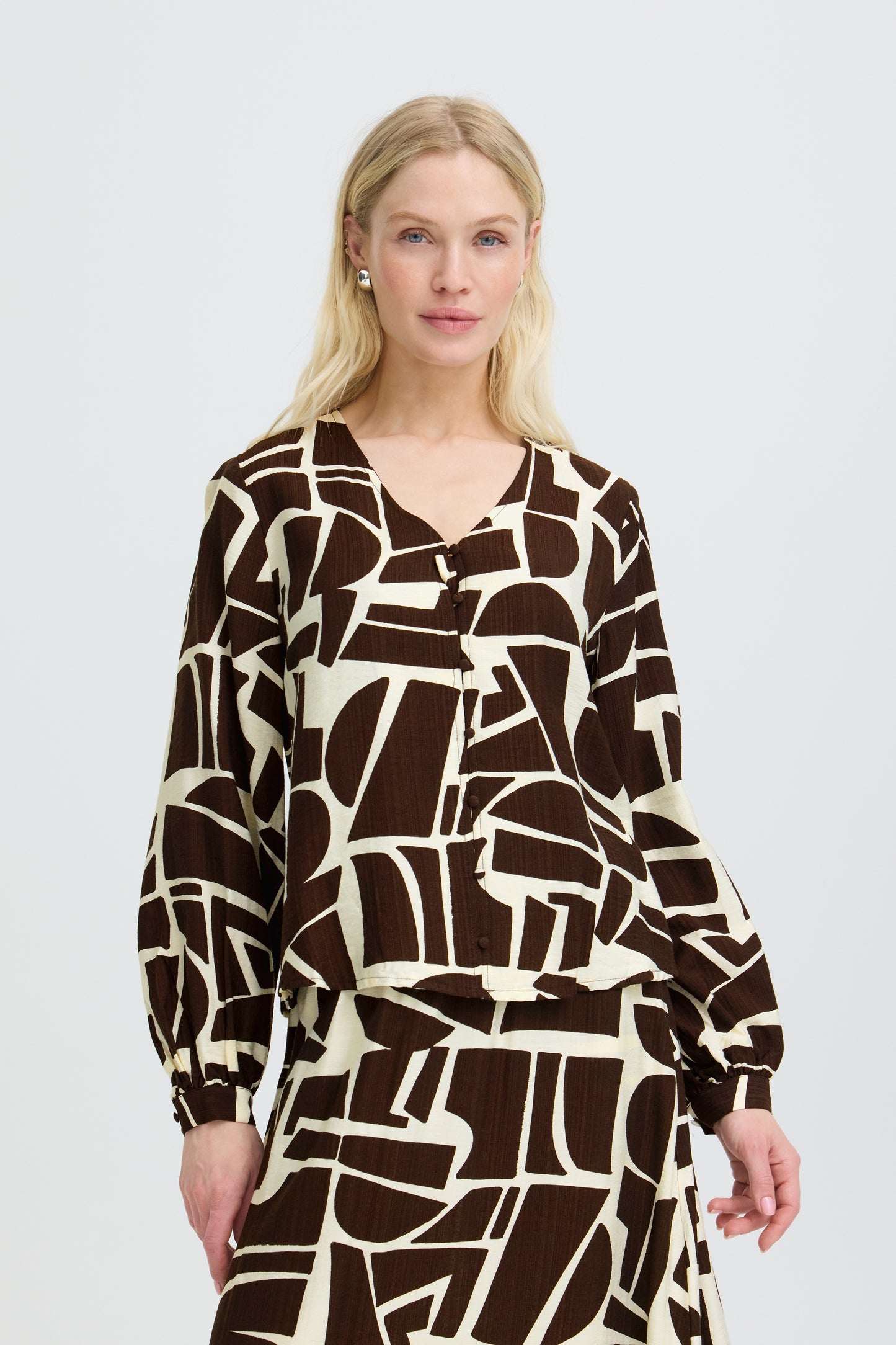 A blonde model wearing a long-sleeve wrap-style blouse with a bold abstract geometric pattern in dark brown and cream, set against a solid light gray background.