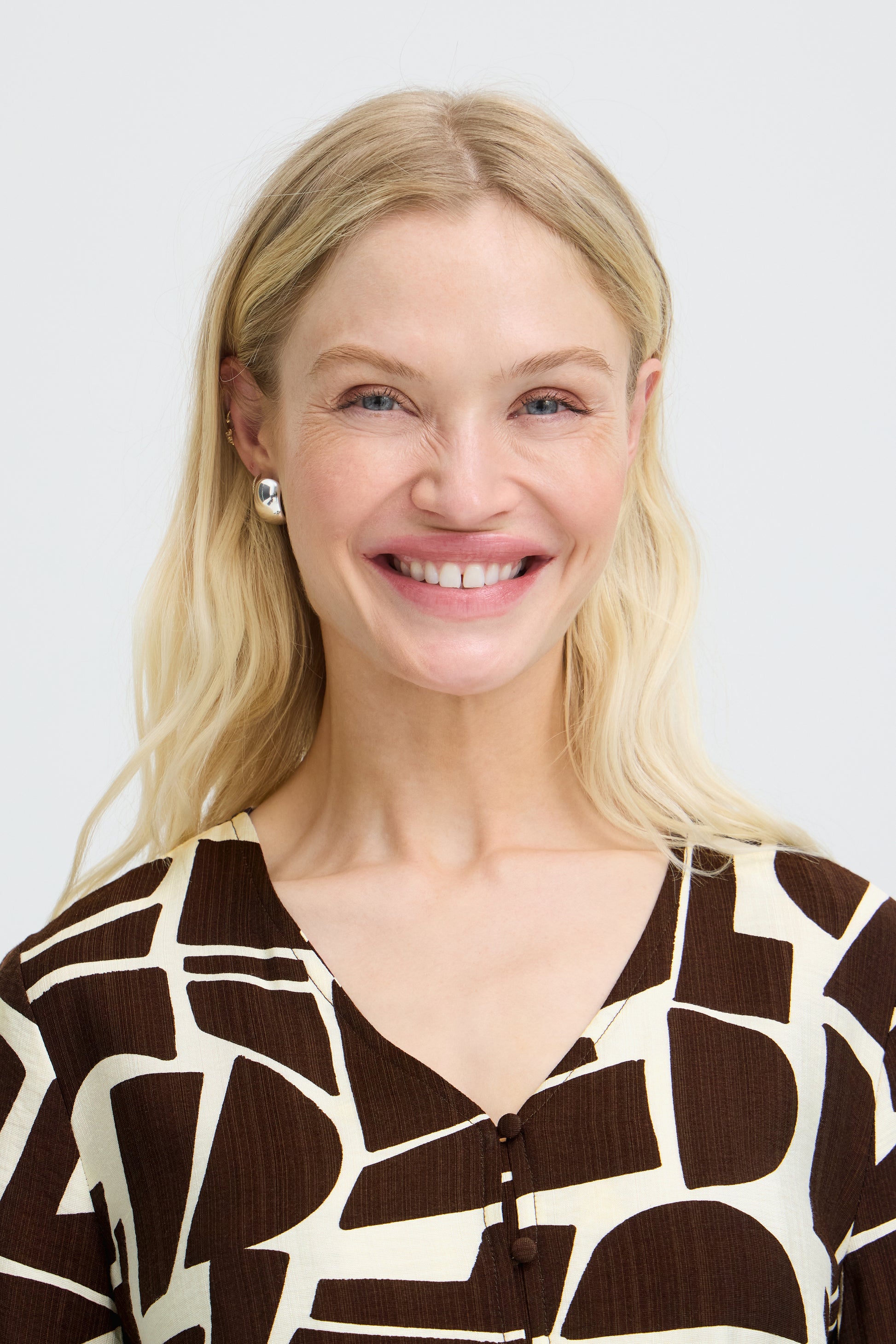 A smiling blonde woman wearing a brown and cream abstract patterned top, photographed against a light gray studio background.