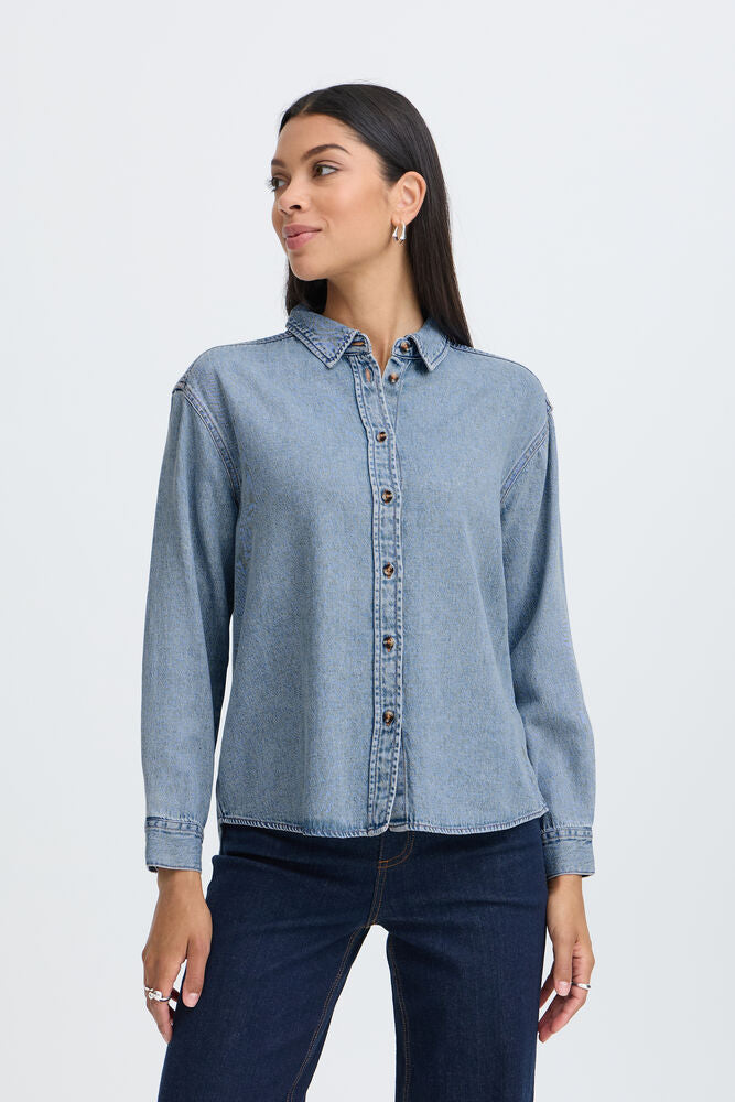 A woman wearing a light blue denim button-up shirt and dark blue jeans, standing against a plain light gray studio background.