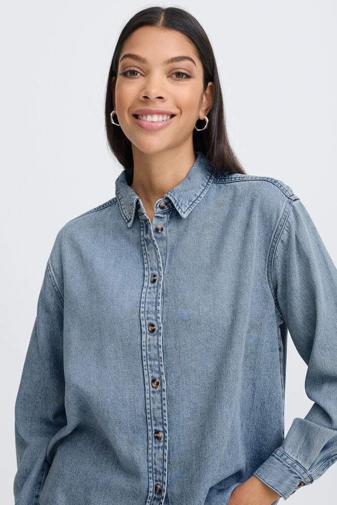 A woman wearing a light blue denim button-down shirt, shown from the chest up against a plain light gray studio background.