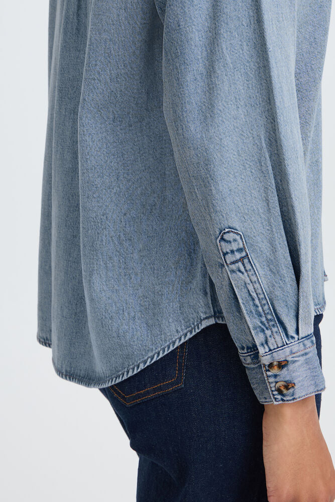 Close-up of a person wearing a light blue denim shirt with dark blue jeans, showing the sleeve cuff with button details and the side pocket of the shirt.