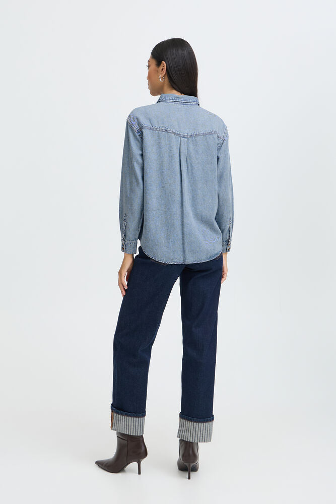 A woman shown from the back wearing a light blue denim button-up shirt, dark blue jeans with rolled cuffs, and brown heeled ankle boots, photographed against a clean white studio background.