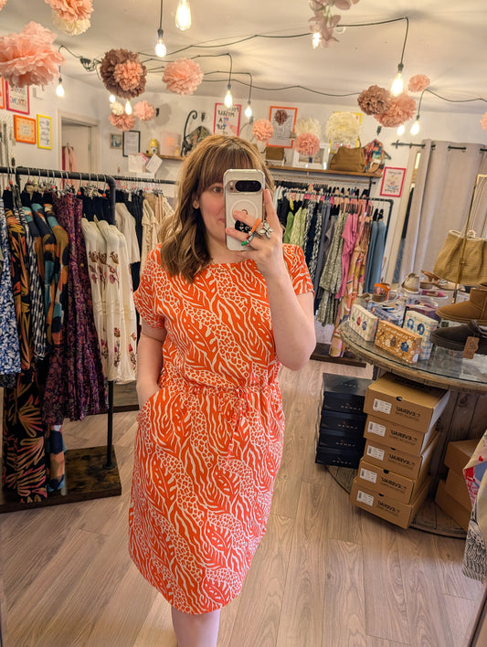 Woman taking a mirror selfie wearing an orange patterned dress in a clothing store.