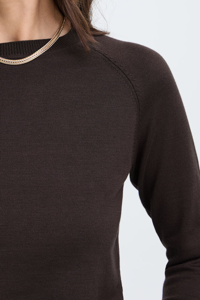 Close-up of a person wearing a dark brown ribbed knit long-sleeve top with a thin gold chain necklace at the neckline, against a light gray background.