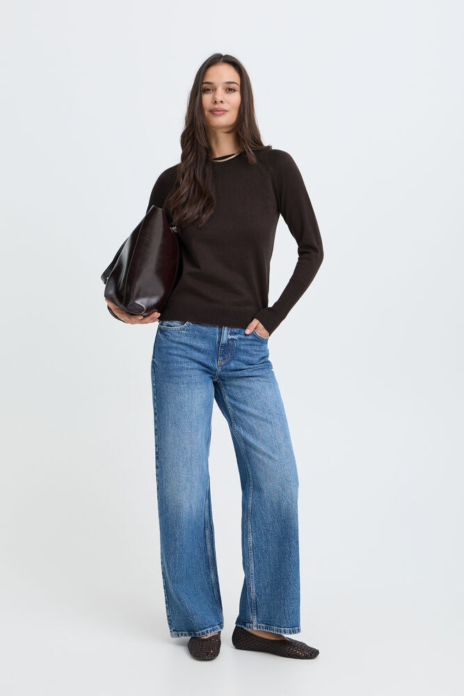 A woman modeling a dark brown long-sleeve knit top, wide-leg blue jeans, and dark brown flats, holding a dark brown leather shoulder bag against a light gray studio background.