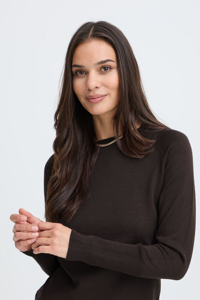 A woman with long dark brown hair wearing a dark brown long-sleeve knit top, photographed against a light gray studio background.