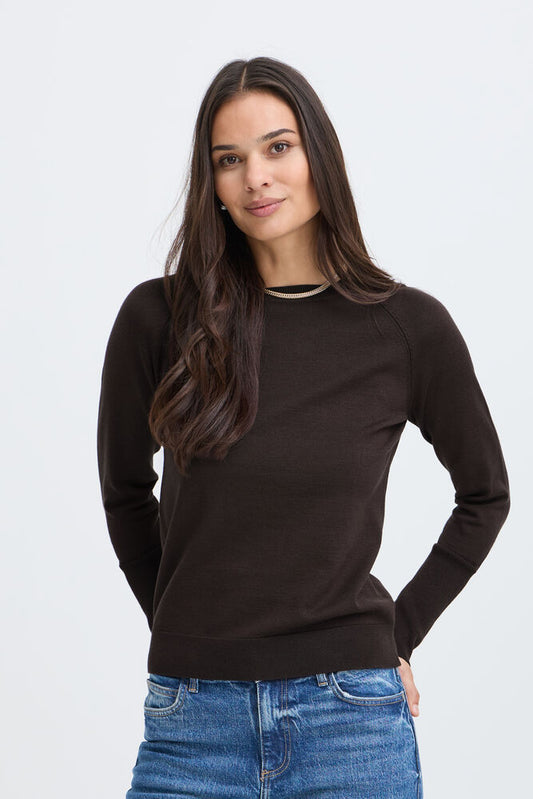 A woman with long dark brown hair wearing a dark brown long-sleeve knit top with a crew neck and blue jeans, standing against a light gray studio background.