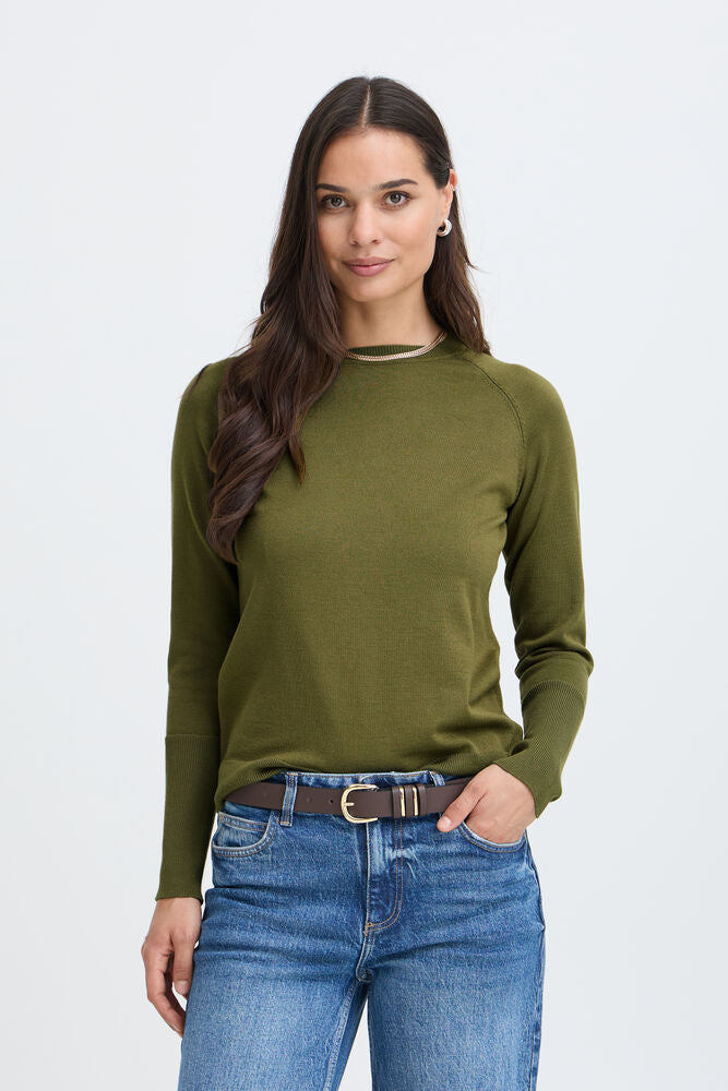 A woman modeling a long-sleeve crewneck knit top in olive green, paired with blue jeans and a brown belt, against a light gray studio background.