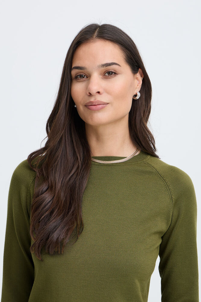A woman with long dark brown hair wearing an olive green long-sleeve knit top with a thin beige trim at the crew neck, photographed against a light gray studio background.