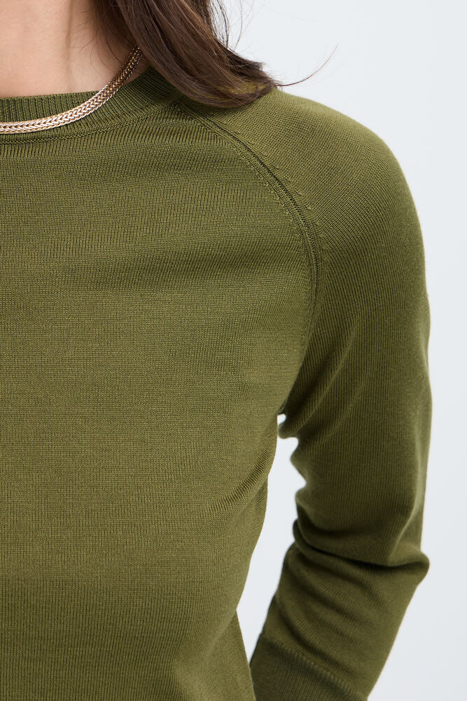 Close-up of a person wearing a ribbed knit crewneck sweater in olive green, showing the shoulder, collar with a thin metallic trim, and part of the arm against a light gray background.