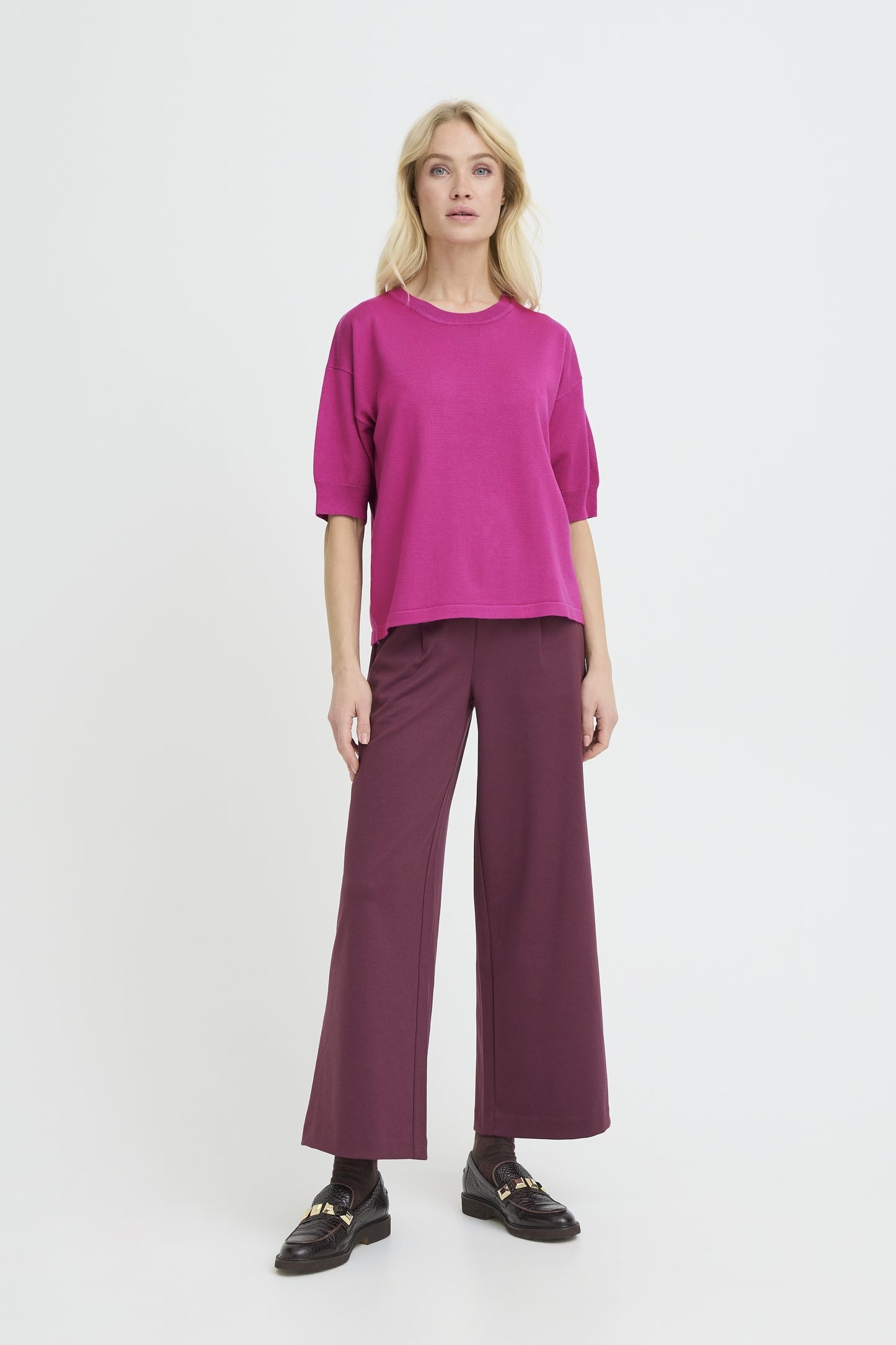 A woman modeling a bright magenta short-sleeve top and dark plum wide-leg trousers with black loafers, standing against a light gray studio background.