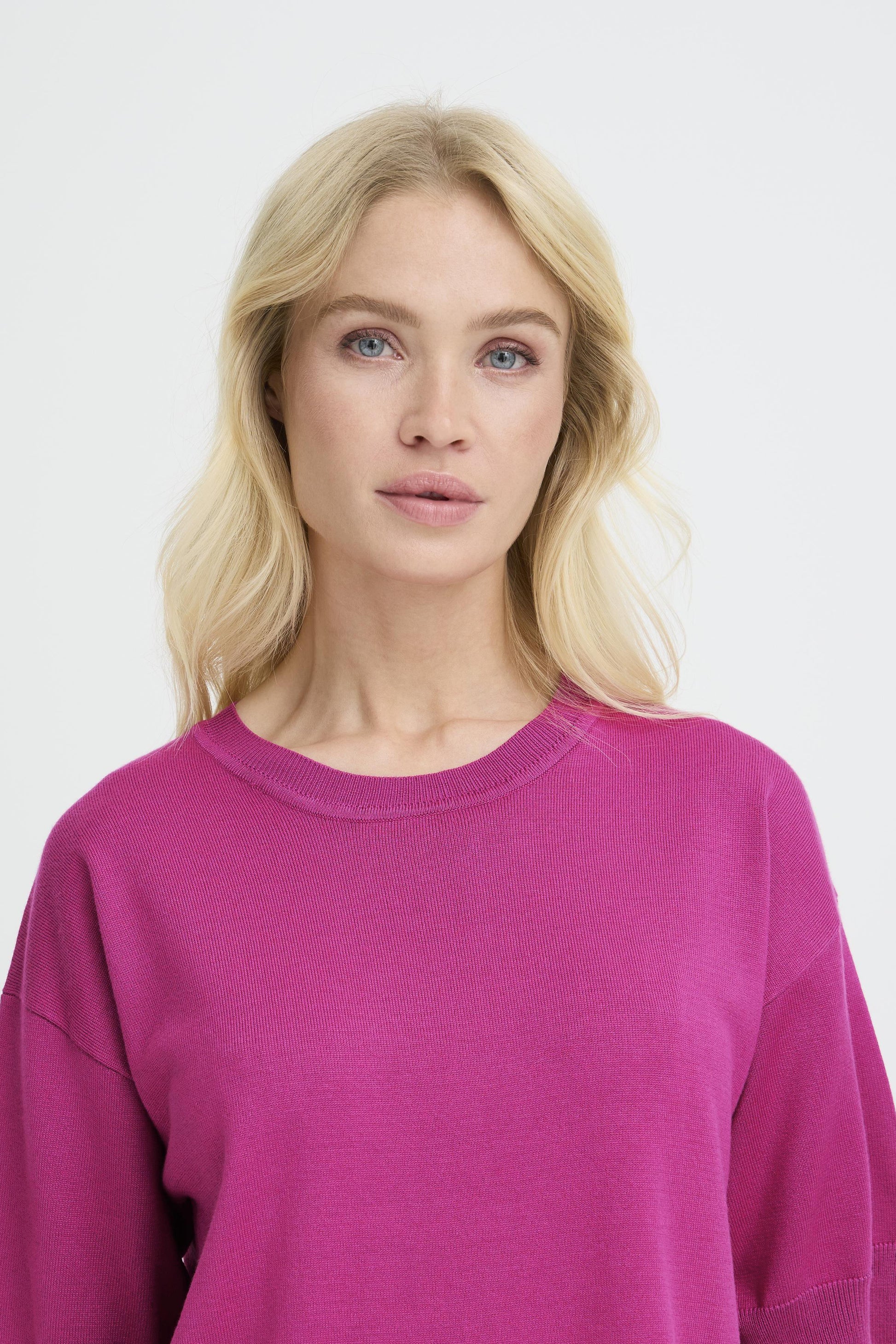 A blonde woman wearing a bright magenta crewneck sweatshirt, shown from the chest up against a plain light gray studio background.