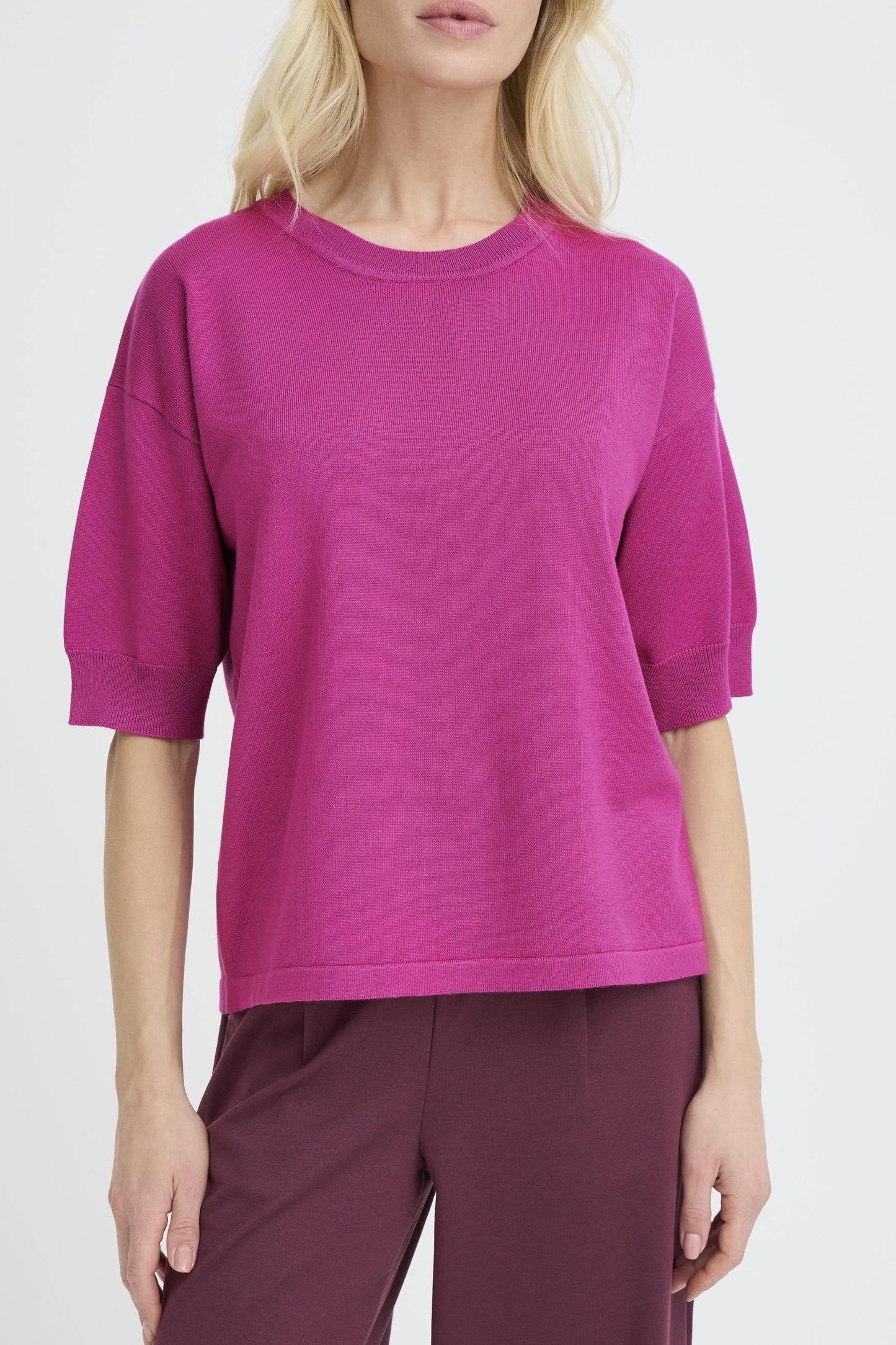 A woman wearing a bright magenta short-sleeve knit top and dark plum pants, cropped from the mouth down against a light gray studio background.