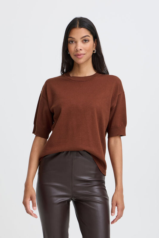 A woman modeling a brown short-sleeve crewneck knit top with gathered sleeves, paired with dark brown faux-leather pants, against a light gray studio background.