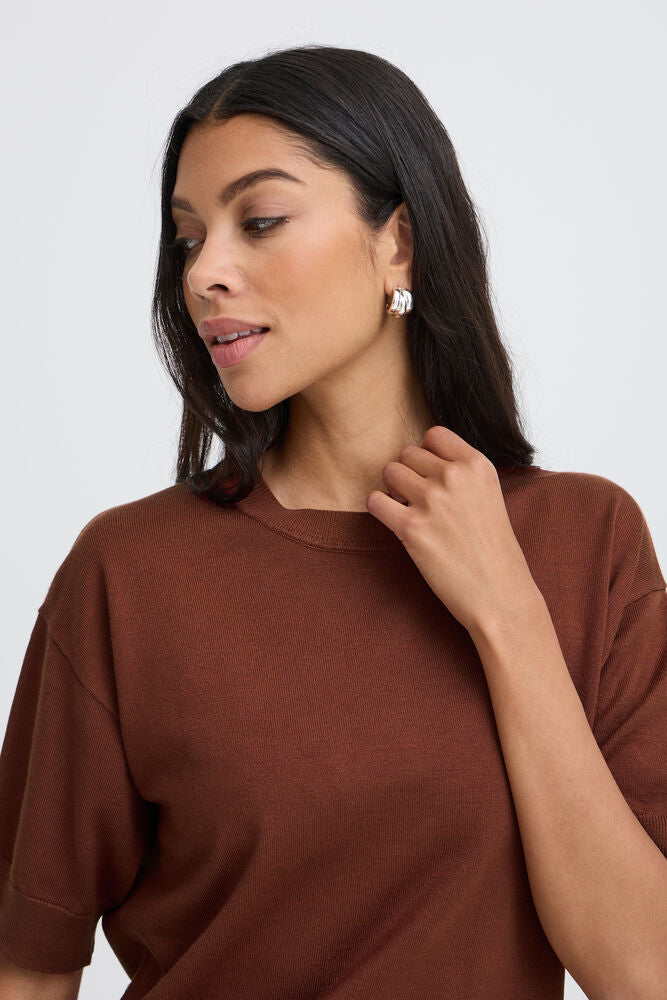 A woman with long dark hair wearing a brown crewneck top and a silver geometric earring, shown in a close-up shot against a light gray background.