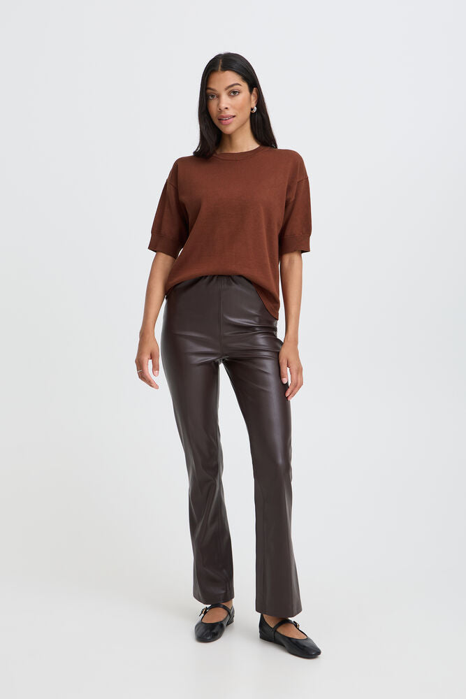 A woman modeling a brown short-sleeve knit top and dark brown faux-leather flare pants with black flats, photographed against a clean light gray studio background.
