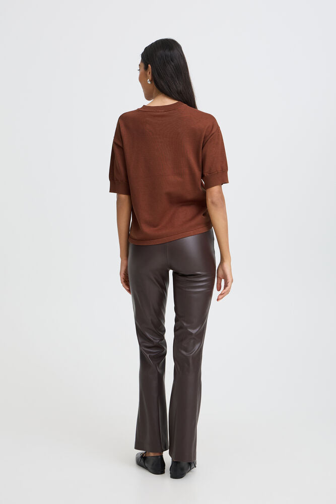 A woman shown from the back wearing a rust-brown short-sleeve knit top and dark brown faux-leather pants, standing against a plain light gray studio background.