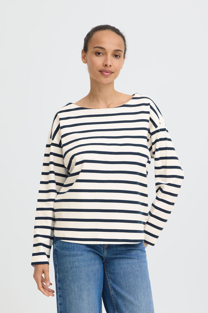 A woman modeling a long-sleeve striped top with a wide boat neckline, featuring horizontal navy and off-white stripes, paired with blue jeans against a light gray studio background.