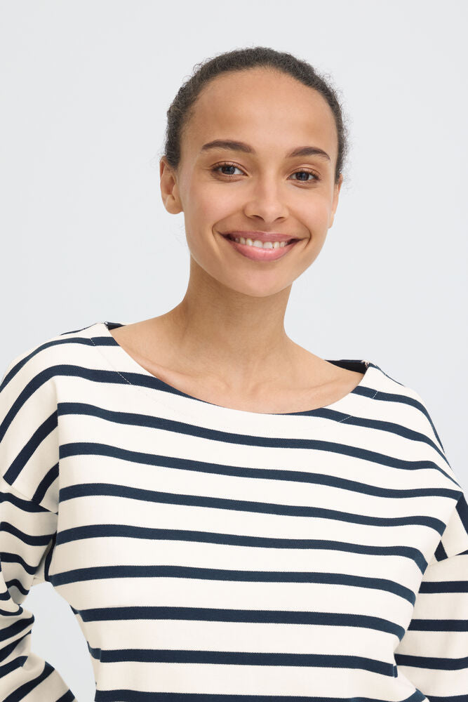 A smiling woman wearing a cream-colored long-sleeve top with navy blue horizontal stripes, photographed against a light gray studio background.