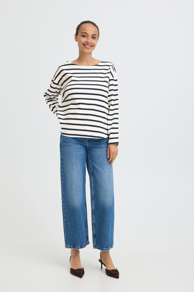 A woman modeling a black and white horizontally striped long-sleeve top with blue wide-leg cropped jeans and dark brown pointed-toe heels, standing against a plain light gray studio background.