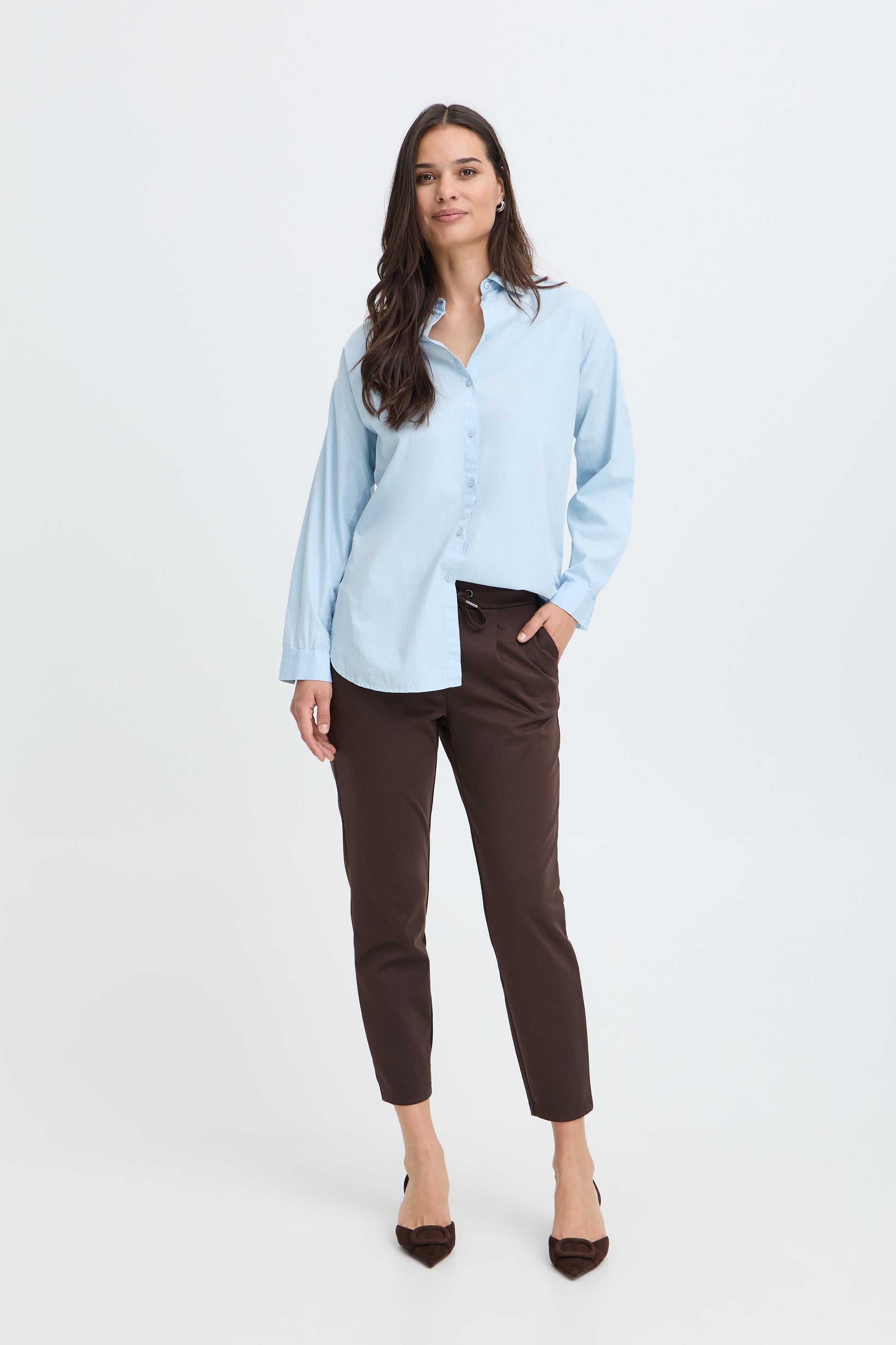 A woman modeling a light blue long-sleeve button-up shirt and dark brown cropped trousers against a plain white studio background.