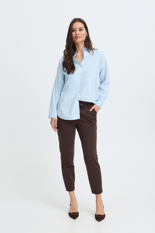 A woman modeling a light blue long-sleeve button-up shirt and dark brown cropped trousers against a plain white studio background.