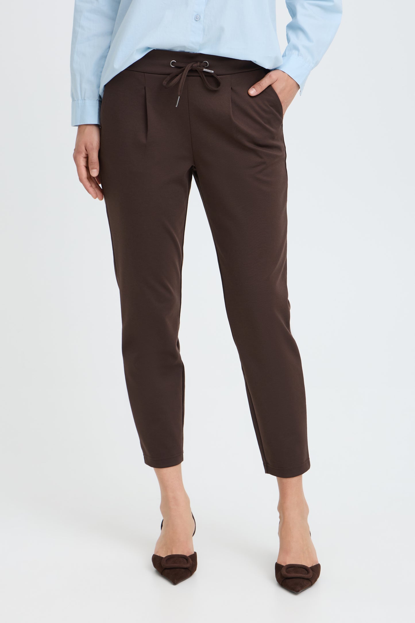 A model wearing dark brown cropped drawstring trousers, styled with a light blue shirt and brown pointed-toe heels against a plain white studio background.