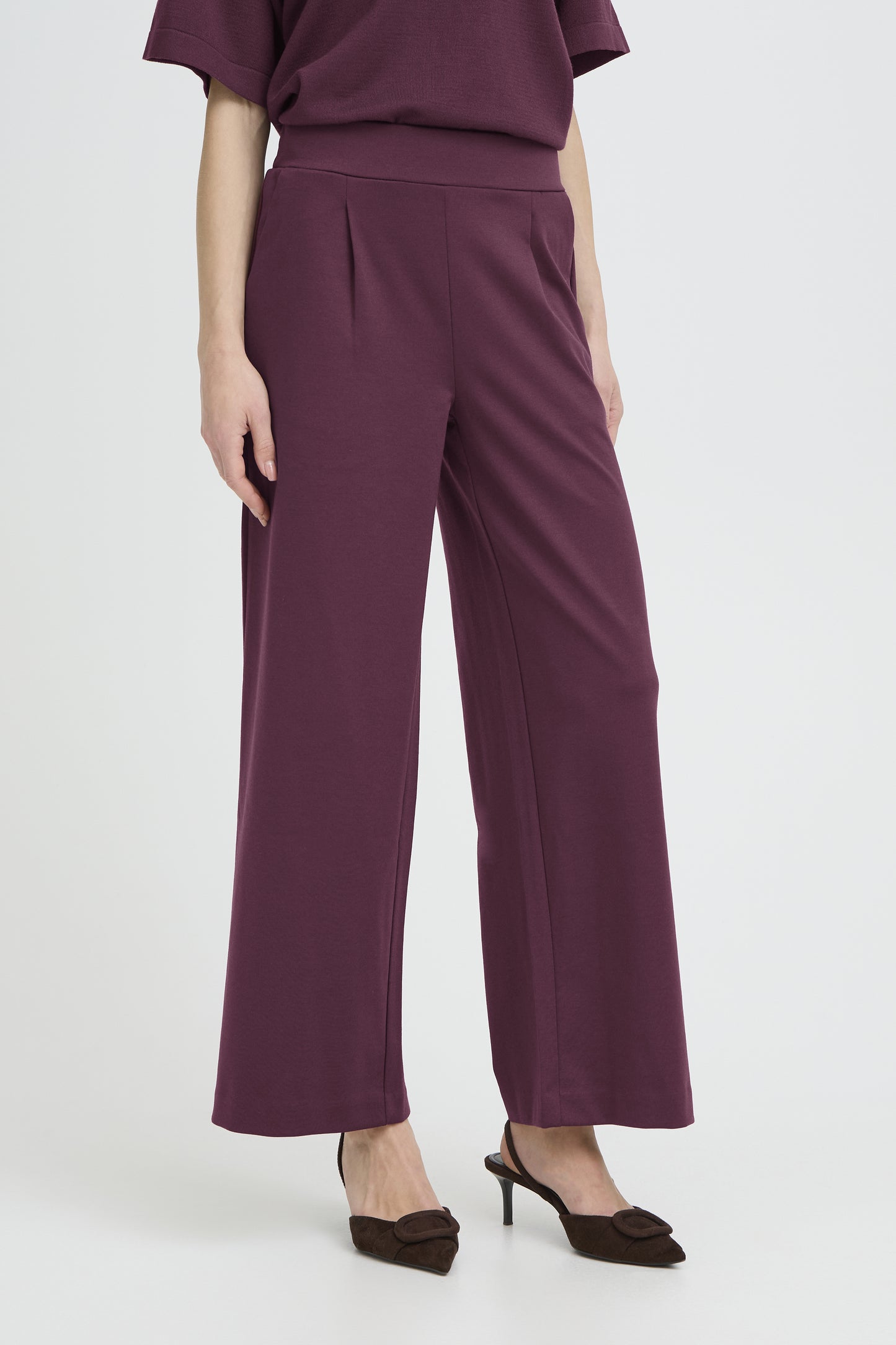 A model wearing a pair of high-waisted, wide-leg trousers in a deep plum purple color, paired with a matching top and dark brown pointed-toe heels, set against a plain light gray studio background.