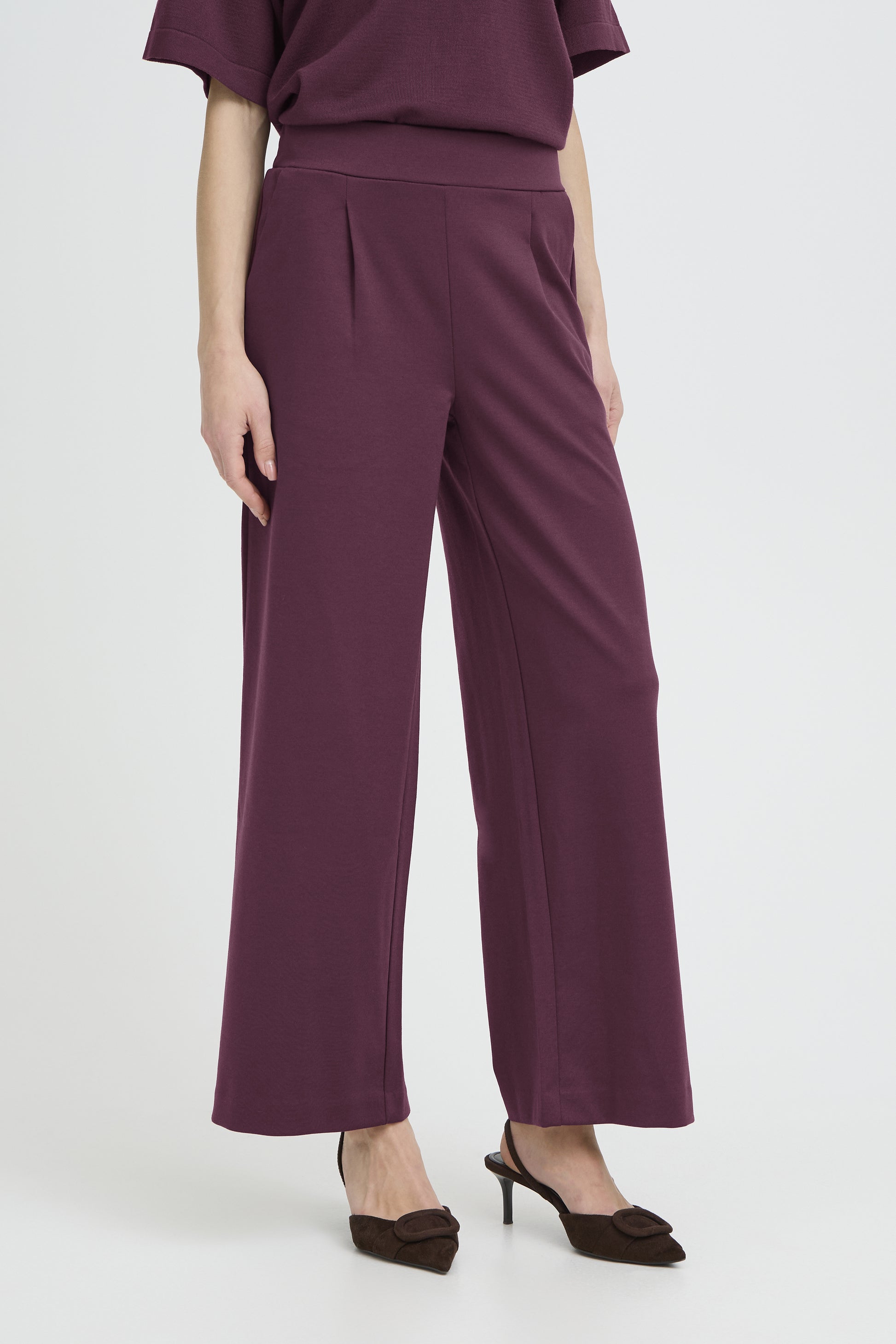 A model wearing a pair of high-waisted, wide-leg trousers in a deep plum purple color, paired with a matching top and dark brown pointed-toe heels, set against a plain light gray studio background.