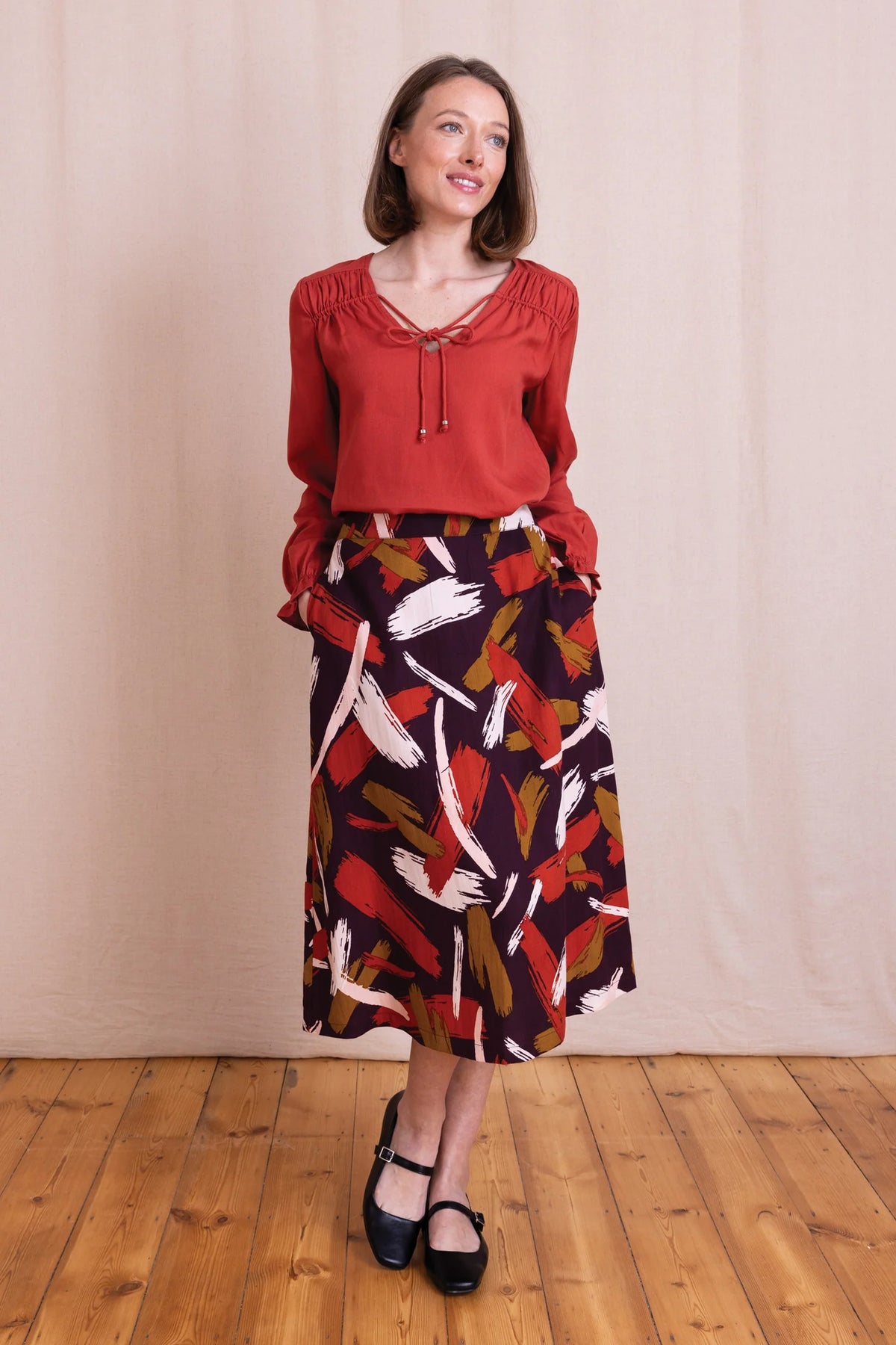 A woman modeling a rust red long-sleeve blouse with a tie neckline and a dark midi skirt featuring a colorful abstract feather print, standing on a wooden floor against a beige fabric backdrop.