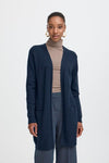 Woman wearing a navy cardigan over a beige turtleneck and gray pants on a white background