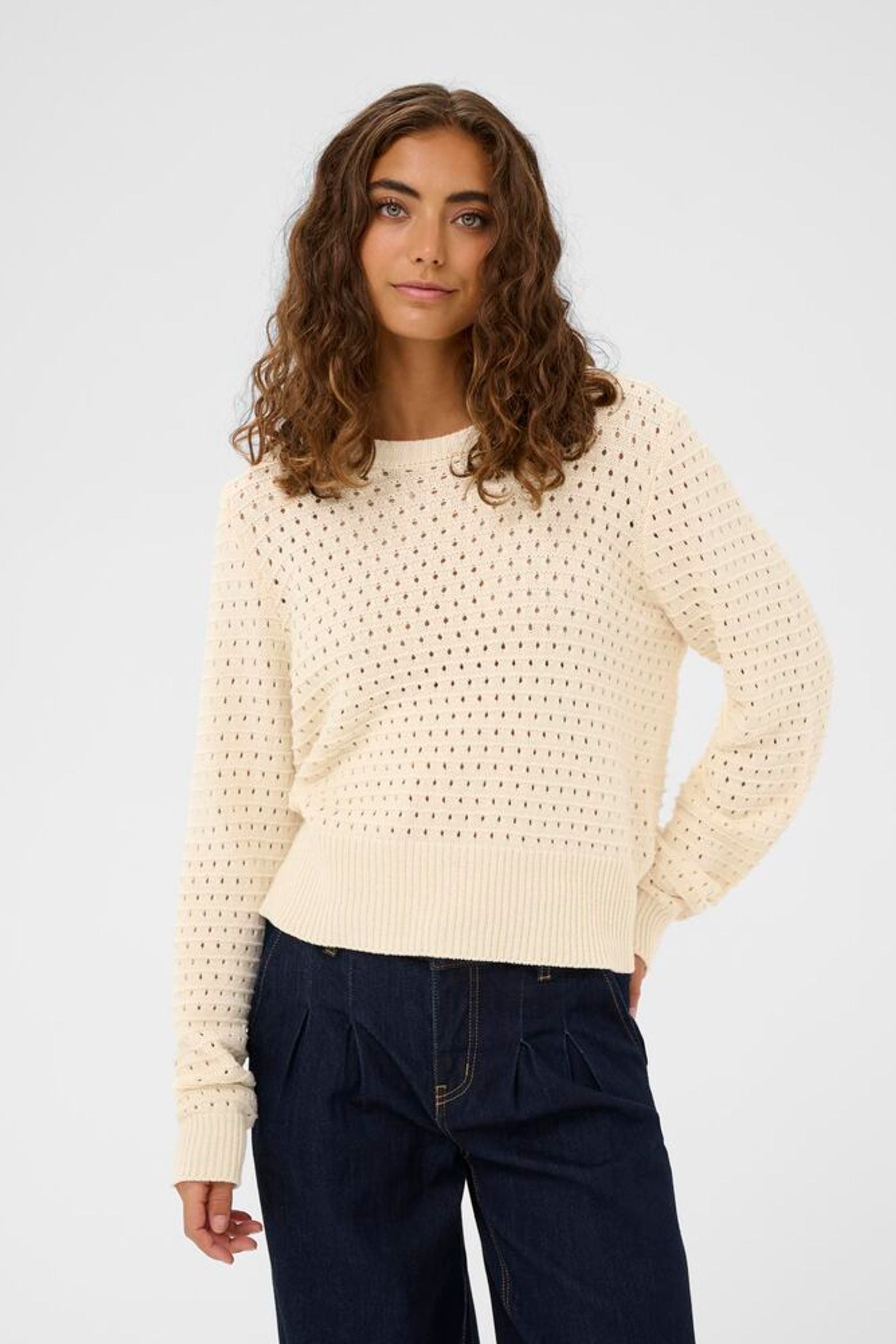 Woman wearing a cream knitted sweater and dark pants on a light gray background