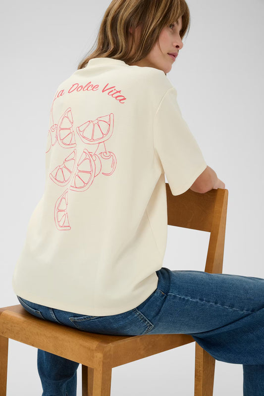 Person wearing a cream-colored t-shirt with red text and graphics, sitting on a wooden chair.