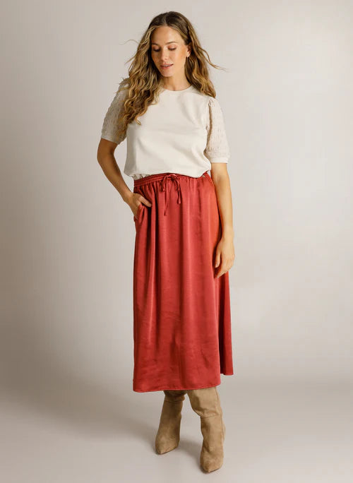A woman modeling a rust-red midi skirt with a drawstring waist, paired with a cream short-sleeve top and beige boots, against a plain light gray studio background.