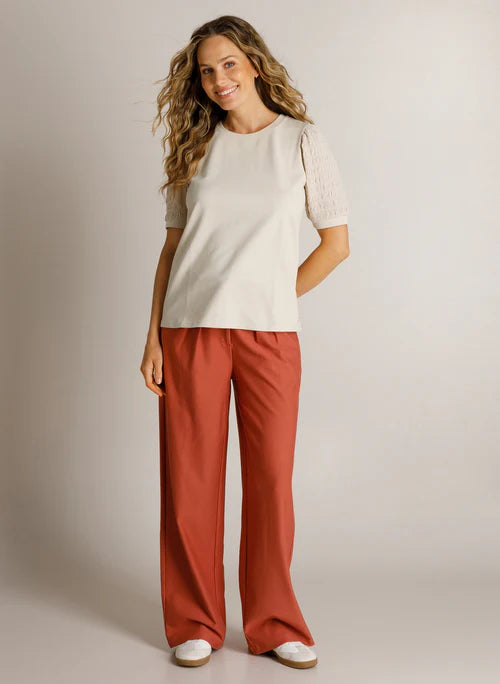 A woman modeling a short-sleeve cream top with textured lace sleeves and wide-leg rust-orange pants, standing against a plain light gray studio background.