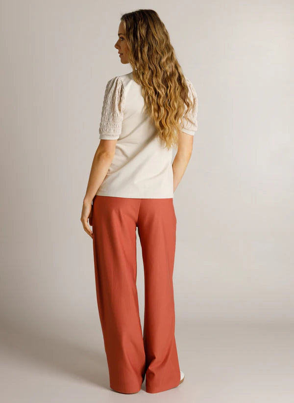 A woman with long wavy light brown hair is shown from the back, wearing a cream short-sleeve top with textured lace sleeves and wide-leg rust-orange trousers, set against a plain light gray studio background.
