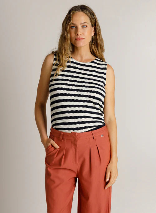 A woman modelling a sleeveless knit top with navy & cream horizontal stripes, paired with rust-coloured pleated trousers, against a plain light grey studio background.