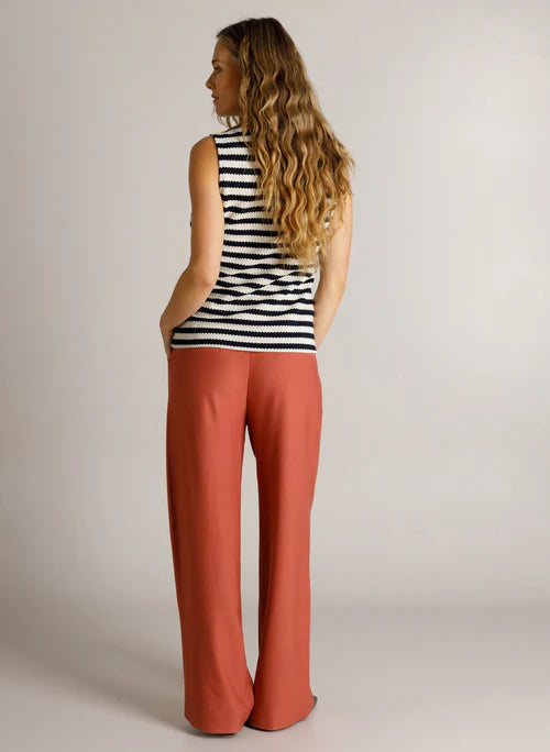 A woman with long wavy blonde hair is shown from the back wearing a sleeveless navy & cream striped top and rust-coloured wide-leg pants against a plain light grey studio background.