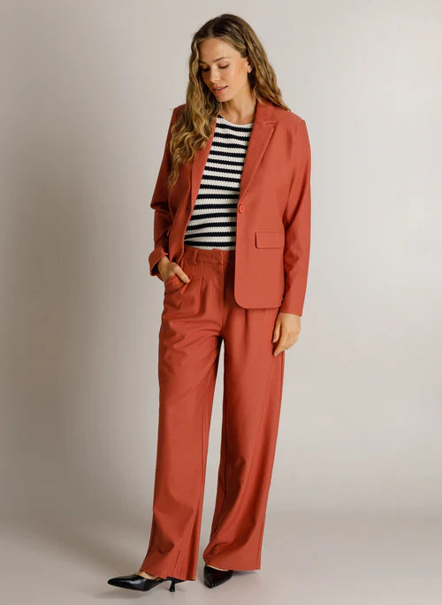A woman modelilng a rust-orange tailored blazer and matching wide-leg trousers, styled with a navy and cream striped top and black pointed heels against a light gray studio background.