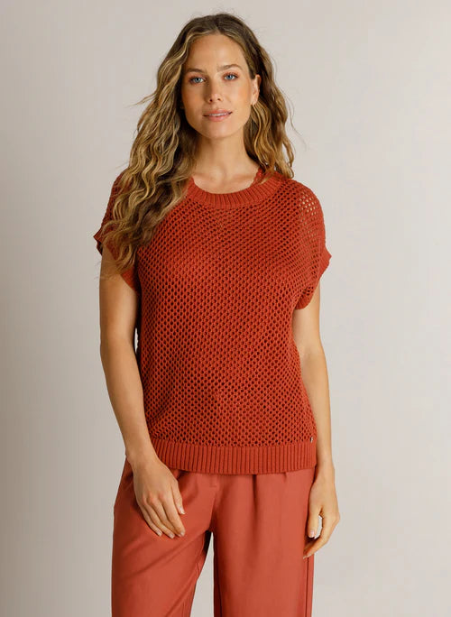 A woman modeling a rust-orange short-sleeve knit top with a textured openwork pattern and matching rust-orange pants against a plain light gray studio background.